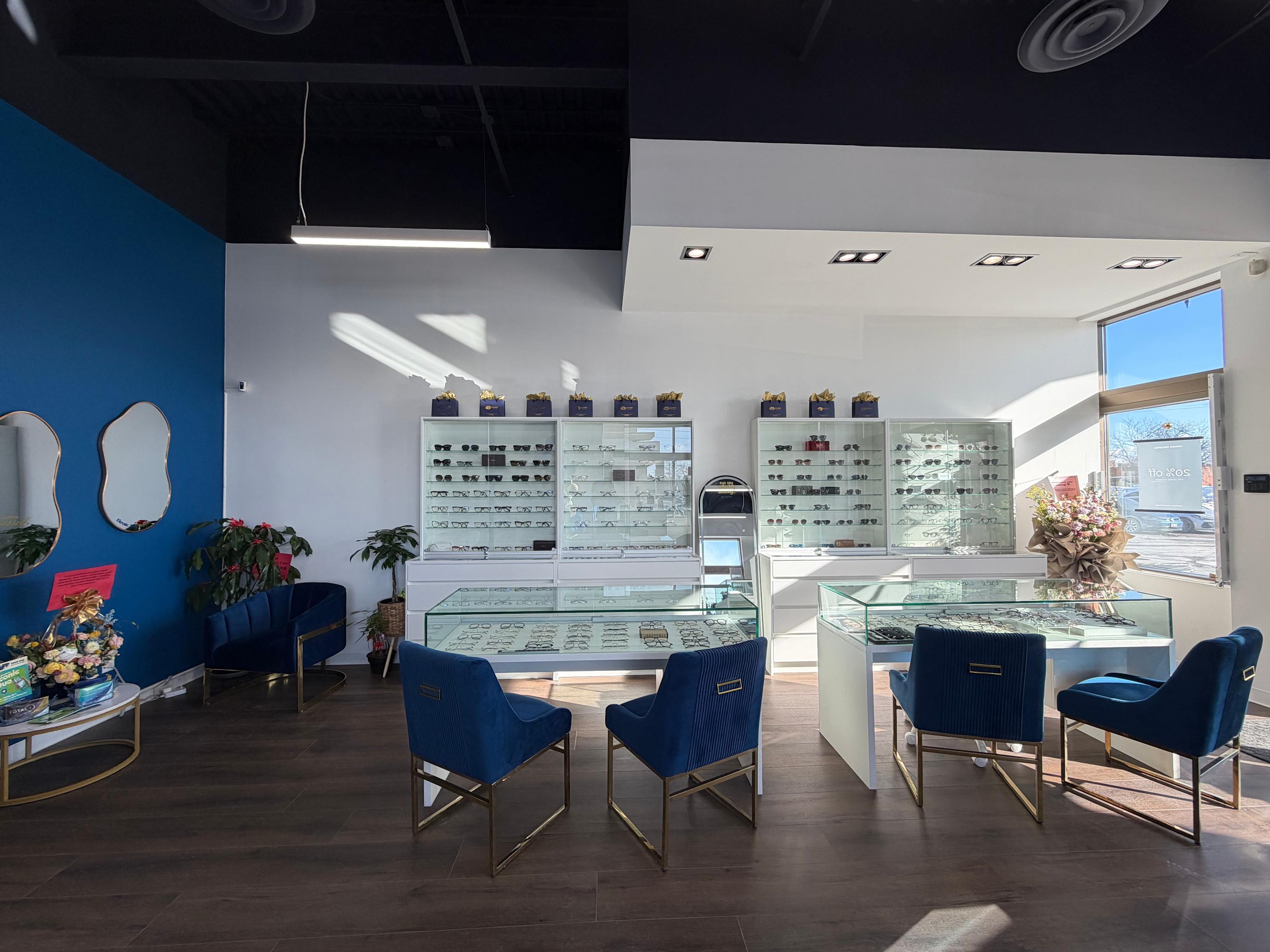 Modern optometry practice interior featuring white display cases filled with eyewear frames, elegant navy blue velvet chairs with gold legs around glass display tables, decorative mirrors on a feature wall painted in rich blue, contemporary linear pendant lighting, recessed ceiling lights, and natural light streaming through large windows with a street view